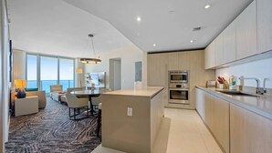A modern hotel room with a dining area, a kitchen, and a large window offering a view of the sea.
