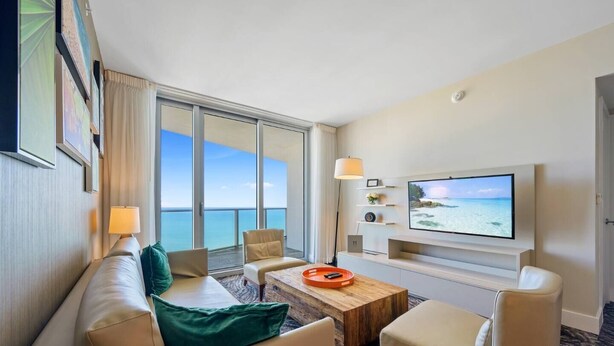 A hotel room with a large window offering a sea view, a flat-screen TV mounted on the wall, a sofa, a coffee table, and a small side table.