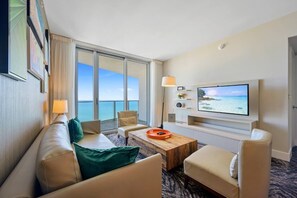 Et hotelværelse med et stort vindue med havudsigt, et fladskærms-tv ophængt på væggen, en sofa, et sofabord og et lille natbord.
