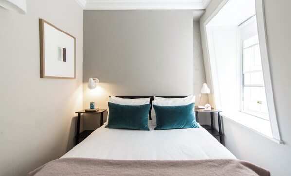 Une chambre d’hôtel bien rangée, avec un lit, deux tables de chevet, un tableau encadré et une fenêtre laissant entrer la lumière naturelle.