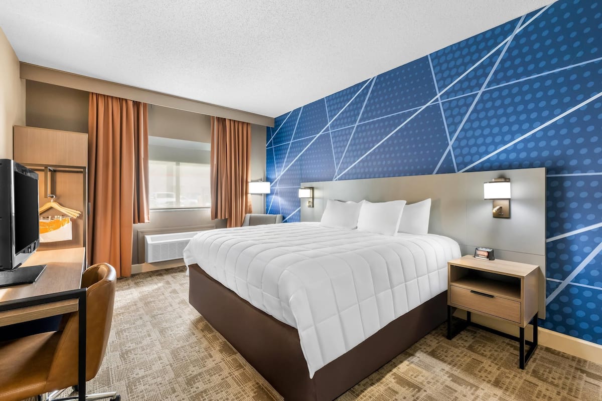 A hotel room with a large bed, a desk with a computer, a chair, a TV, a nightstand, and a wall with a geometric pattern.