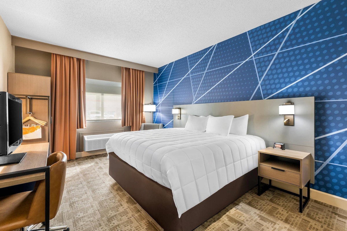 A hotel room with a large bed, a desk with a television, a chair, a nightstand, and a wall with a geometric pattern.