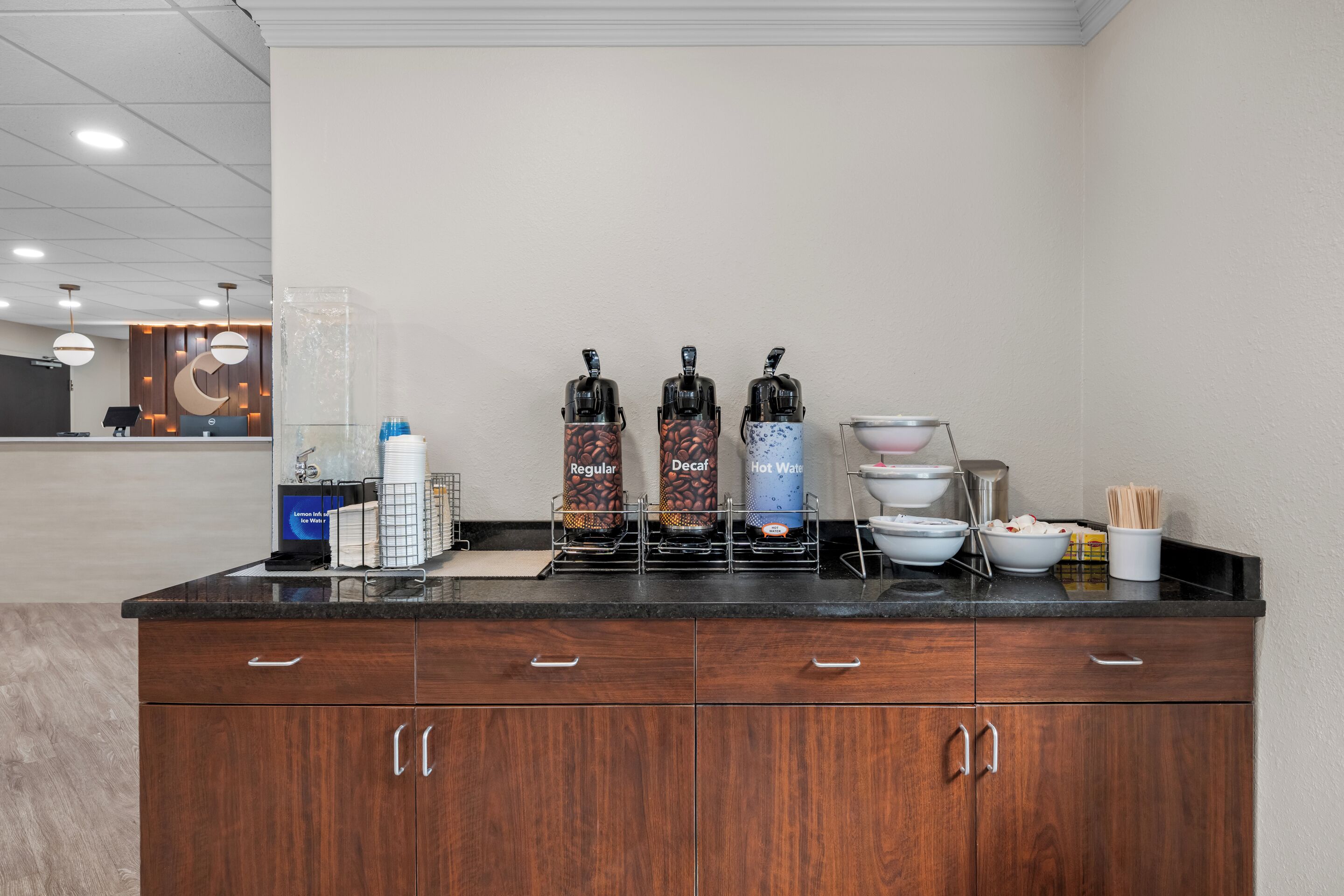 A kitchen area with a coffee station featuring decaf, regular, and hot water options, a water dispenser, and a bowl with utensils.
