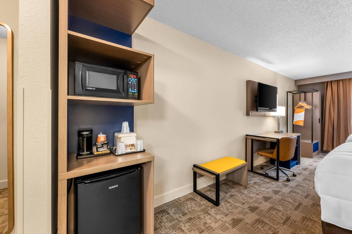 A hotel room with a microwave, coffee maker, mini-fridge, desk, chair, and a bed.