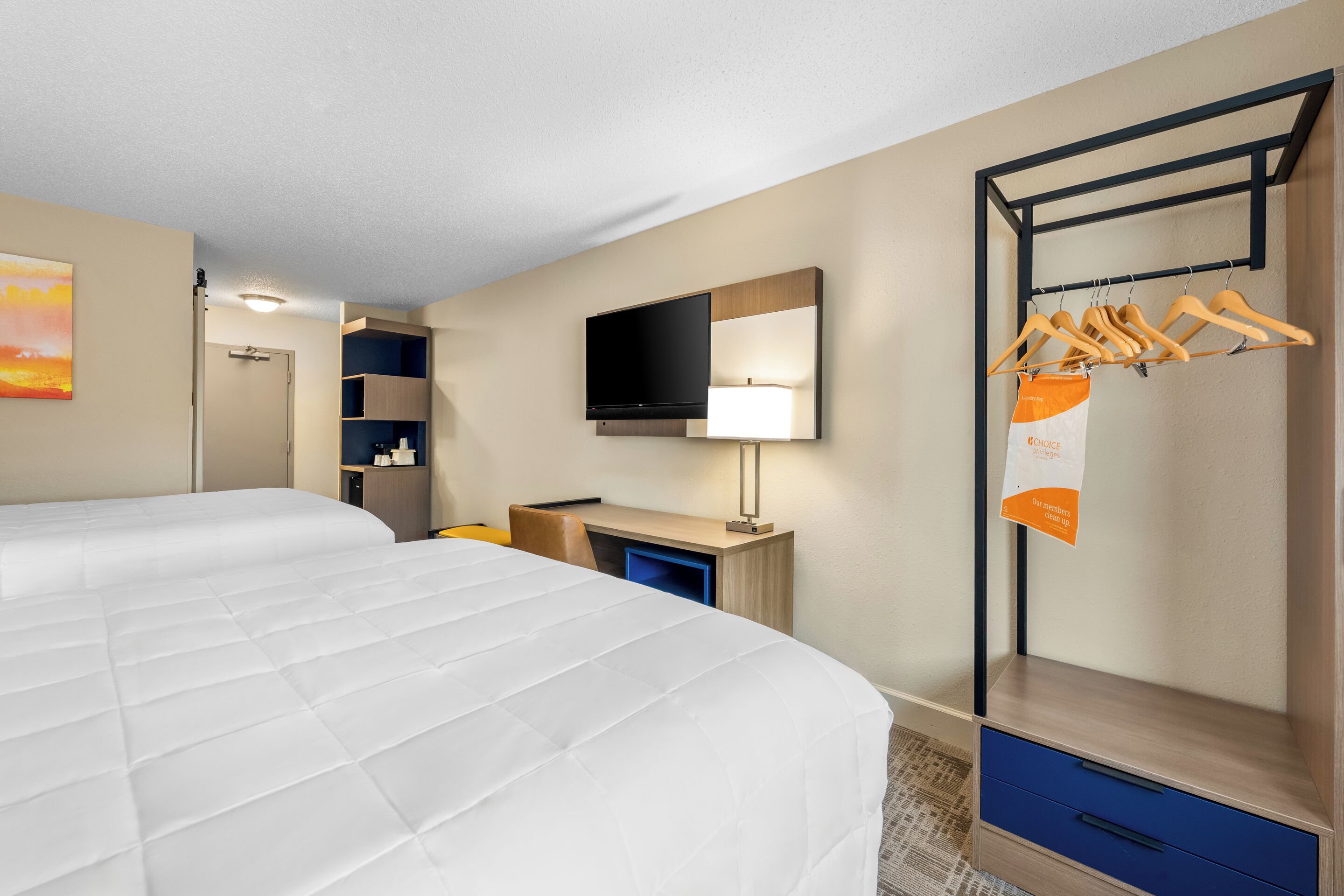 A hotel room with two beds, a desk with a flat-screen TV, a hanging clothes rack, and a wall-mounted TV.