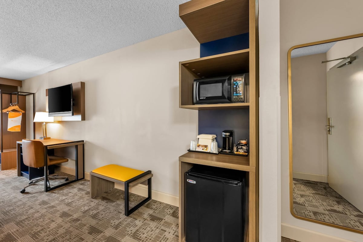 A hotel room with a desk, chair, microwave, coffee maker, and a small refrigerator.