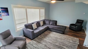 A living room with a sectional sofa, a gray armchair, a black recliner, and a large window with blinds.
