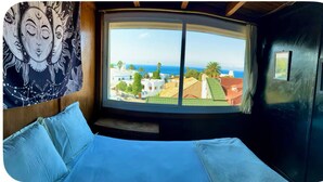 A room with a curved window, a bed with blue bedding, a wall tapestry, and a view of buildings and the sea.