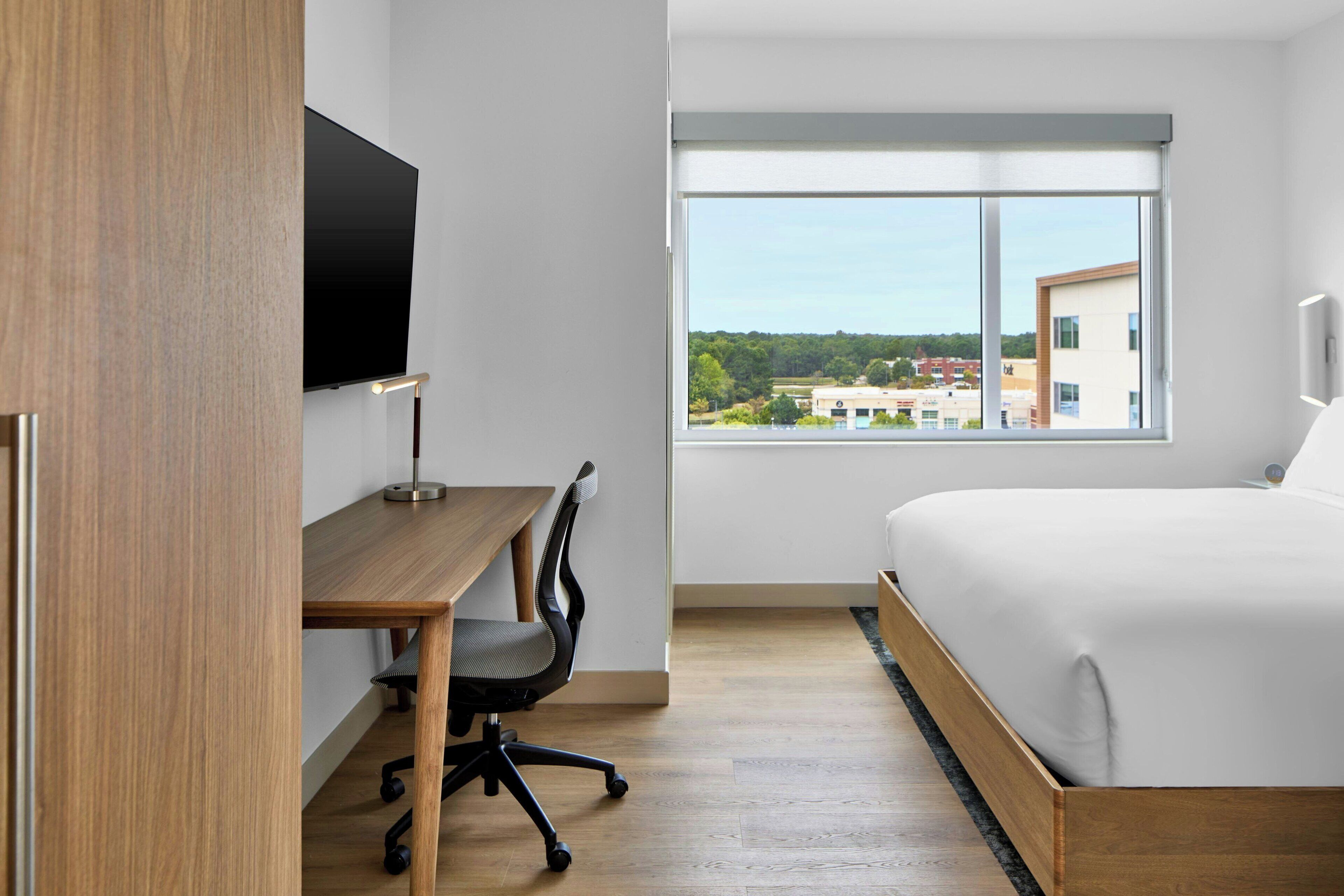 A modern hotel room with a wooden desk, a black office chair, a large bed with white bedding, and a large window offering a city view.