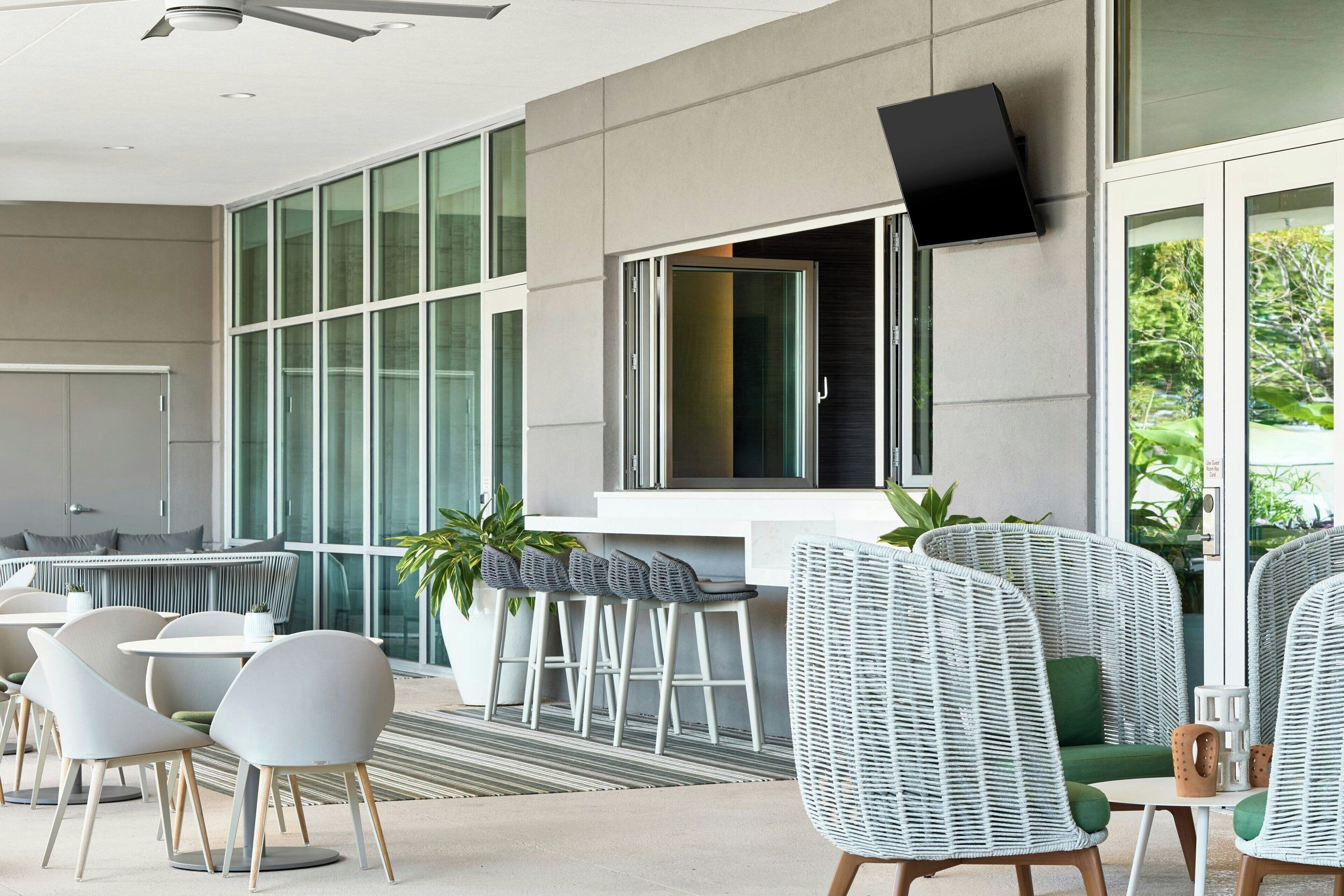 Un espace extérieur moderne, aménagé avec des chaises et des tables en osier, un coin bar et une grande fenêtre offrant une vue sur la verdure.