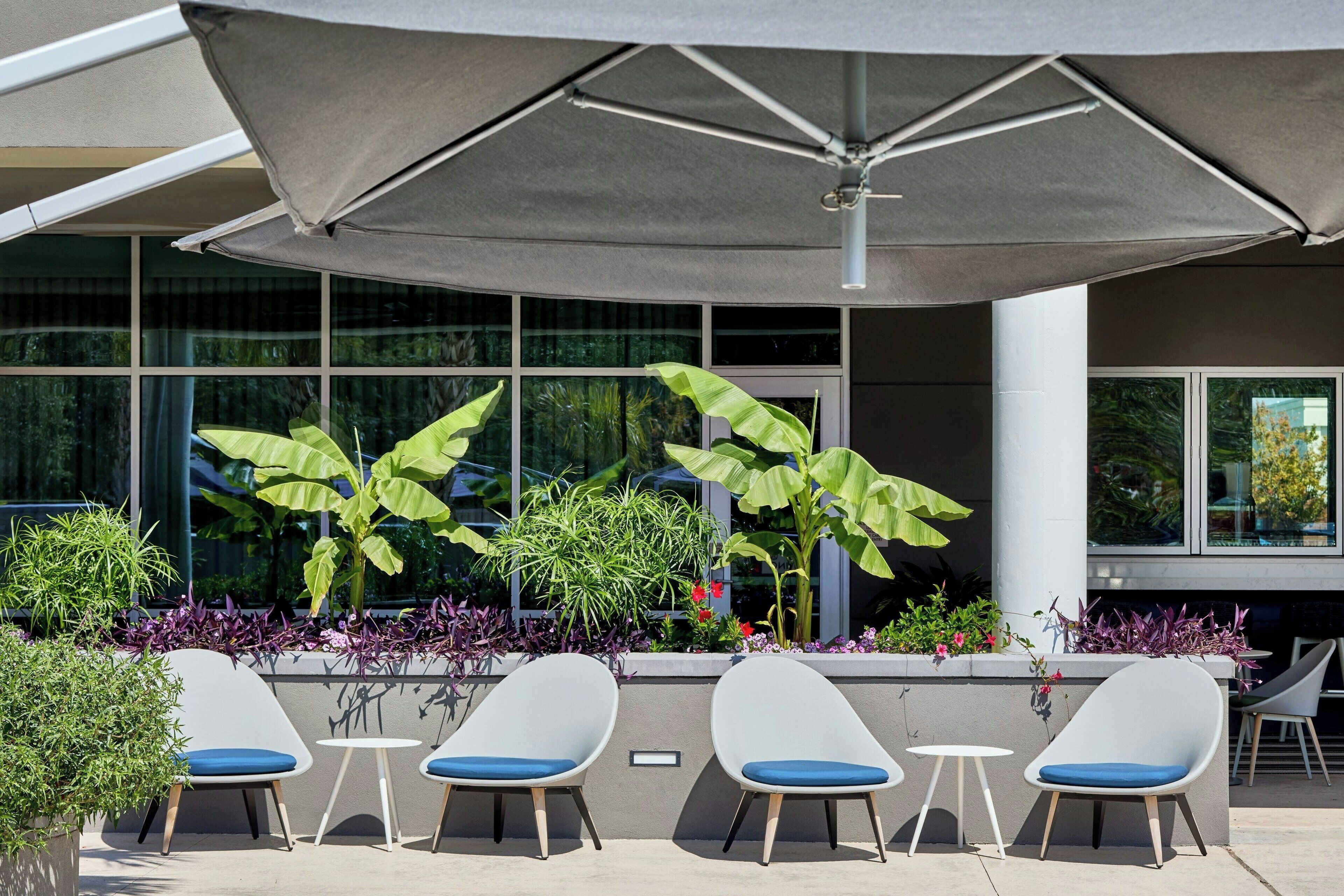 A seating area with four white chairs and a large umbrella, surrounded by potted plants and a modern building facade.