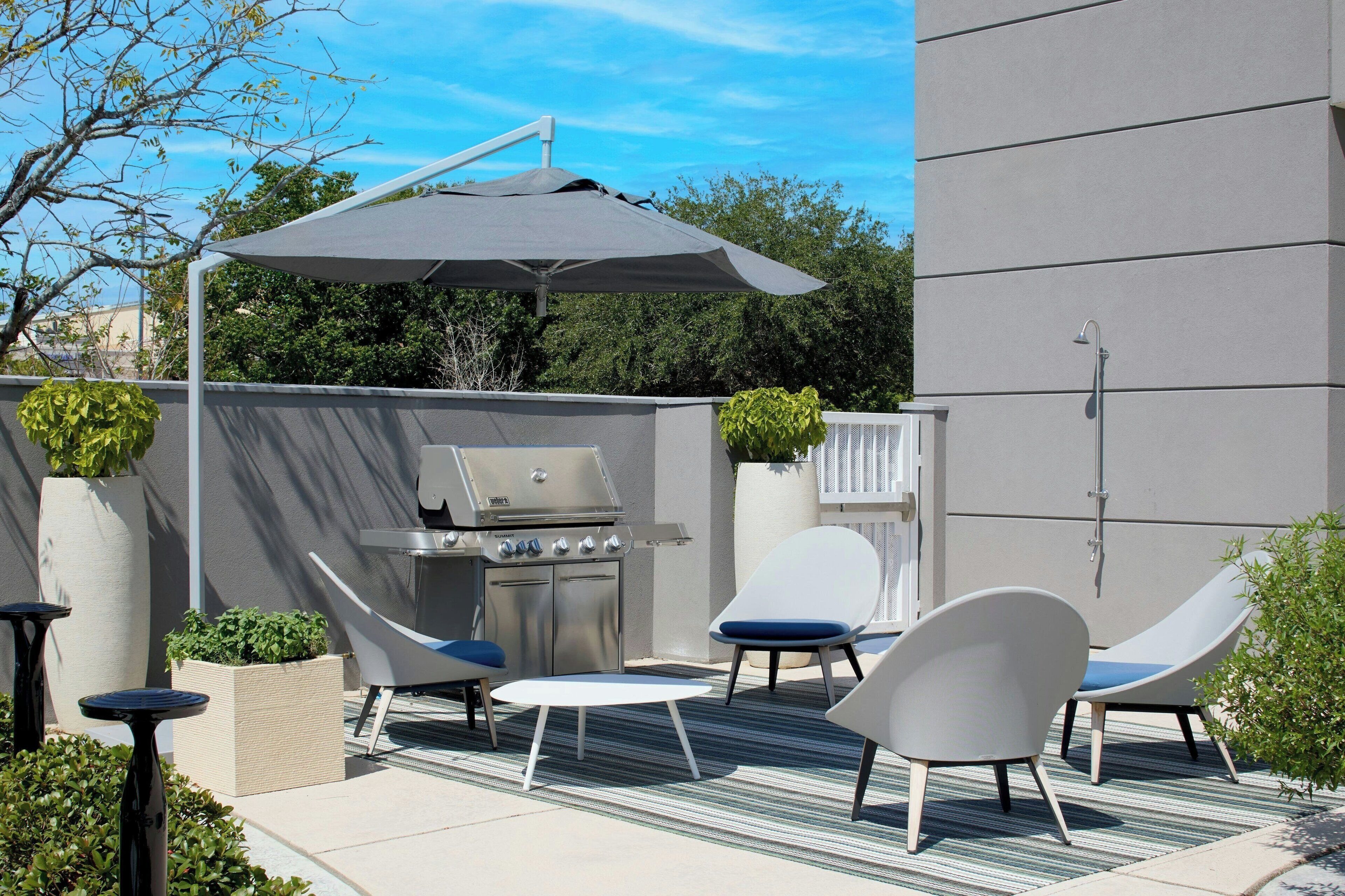 Une terrasse extérieure équipée d’un barbecue, de deux chaises longues, d’une table et d’un grand parasol.