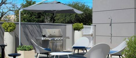 Une terrasse extérieure équipée d’un barbecue, de deux chaises longues, d’une table et d’un grand parasol.