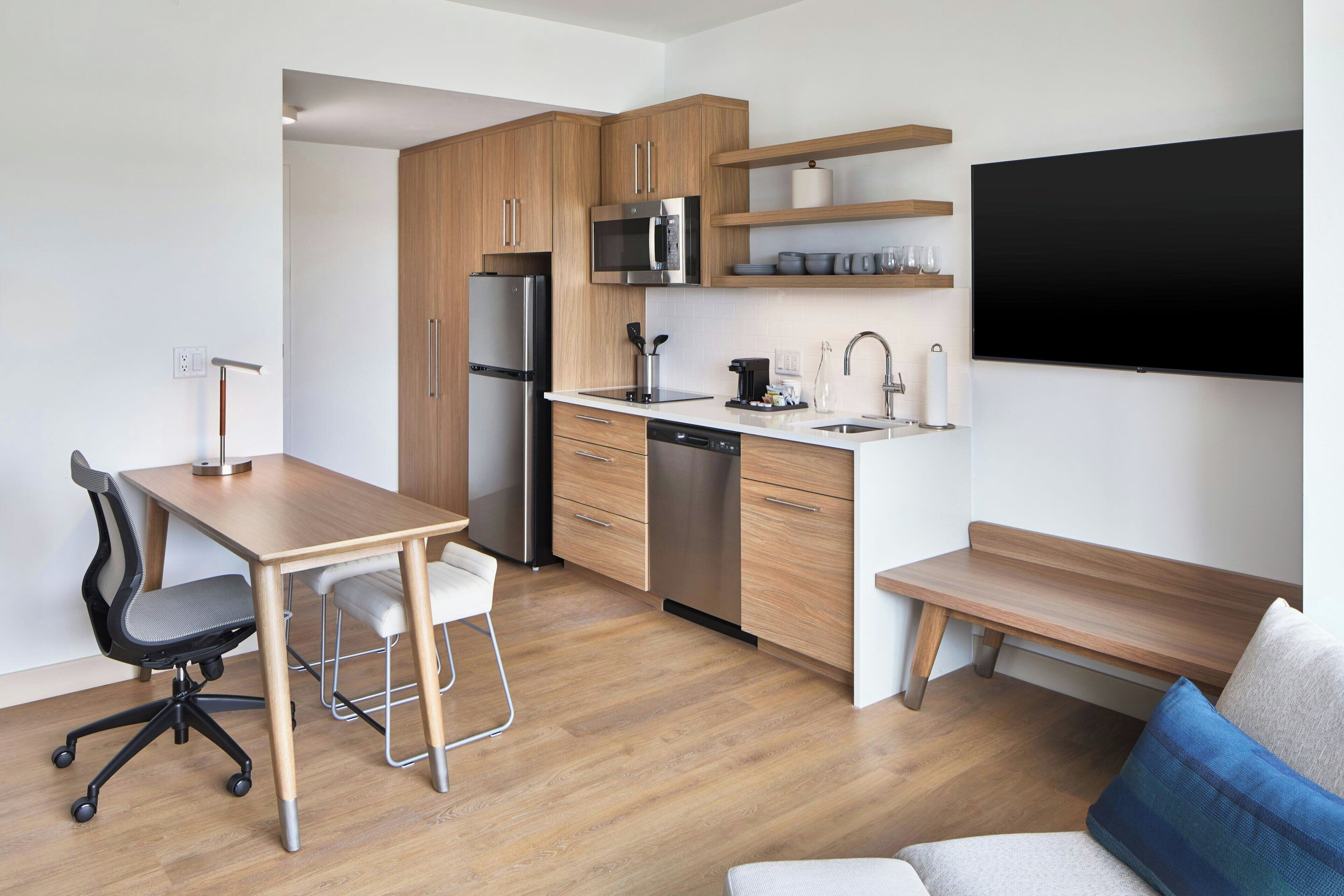A compact kitchen with wooden cabinets, a stainless steel refrigerator, a microwave, and a dishwasher. There is a dining table with two chairs and a bench. A flat-screen TV is mounted on the wall above the bench.