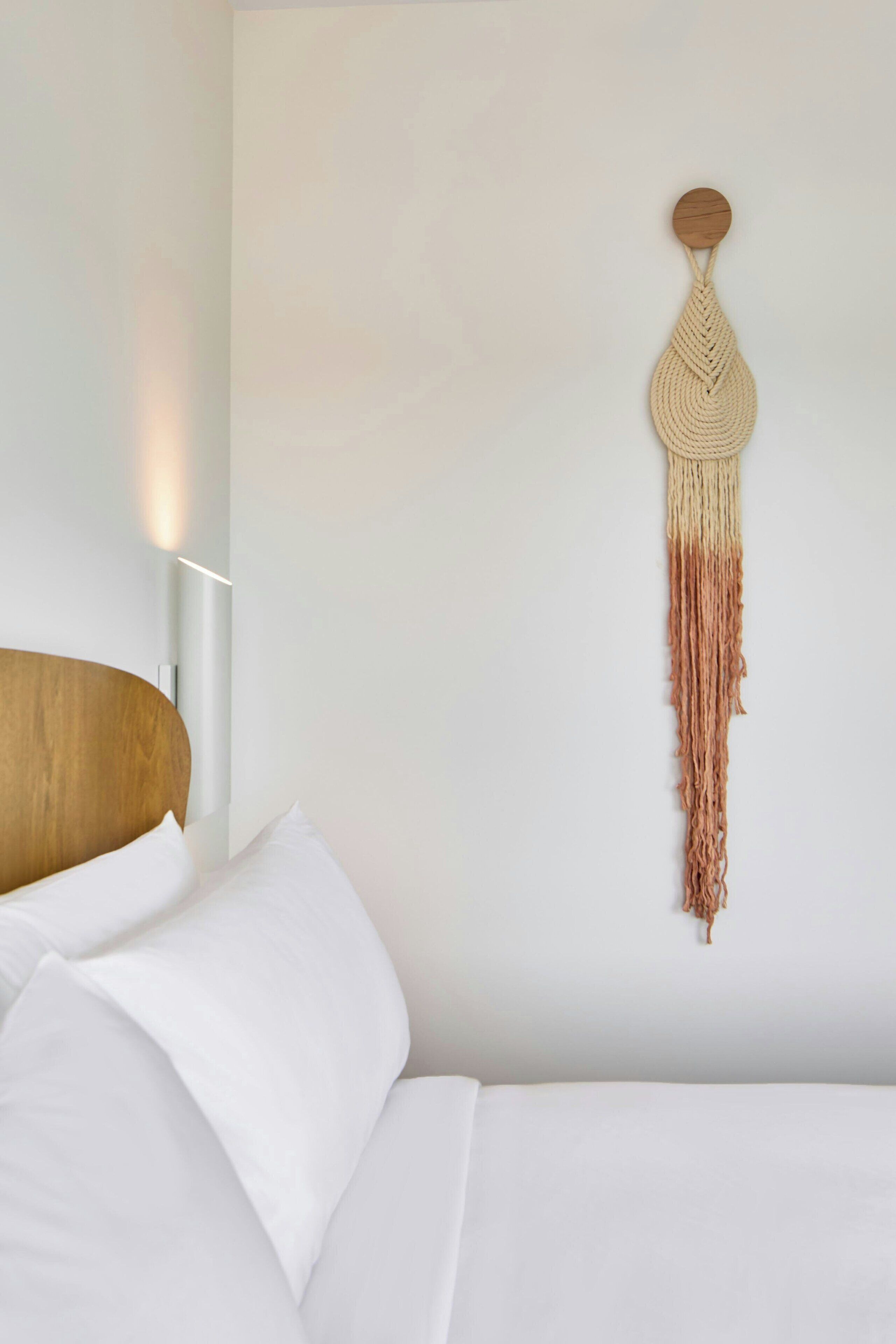 Un lit avec du linge de lit blanc et une tête de lit en bois, une décoration murale en macramé et un luminaire fixé au mur.