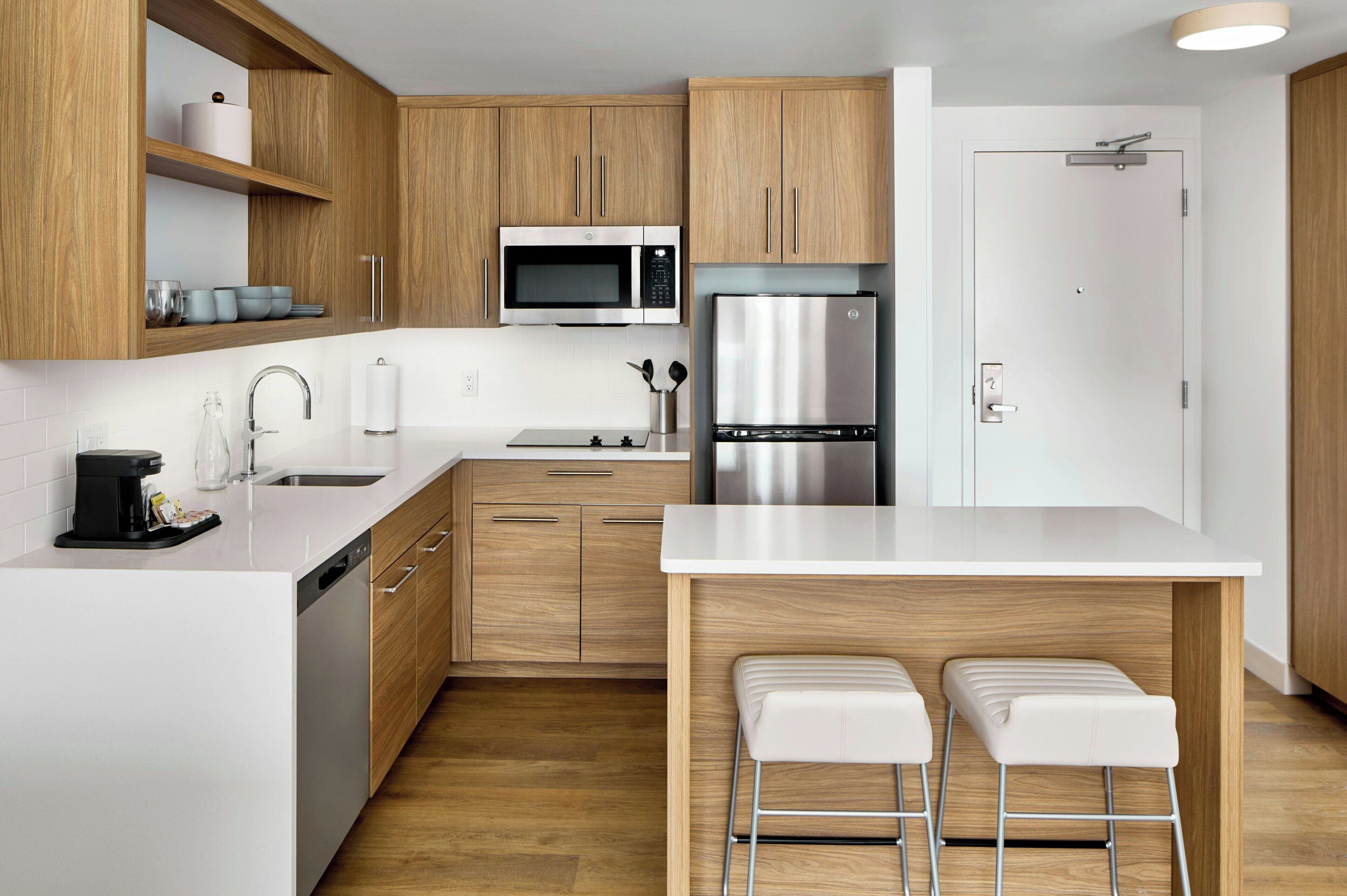 Una cocina moderna con armarios de madera, un refrigerador de acero inoxidable, un microondas y una cafetera. Hay dos taburetes blancos y una encimera blanca.