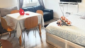 Une pièce de taille modeste comprenant un lit, un téléviseur fixé au mur, une table à manger avec des chaises et une fenêtre avec des rideaux.
