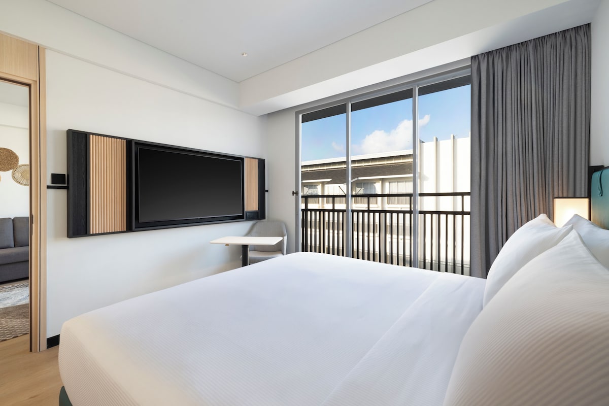 Ein modernes Hotelzimmer mit einem großen Bett, einem flachbildfernseher an der Wand, einem kleinen Schreibtisch und einem Balkon mit Blick auf die Stadt.