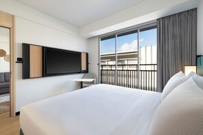 Una habitación de hotel moderna con una cama grande, un televisor de pantalla plana montado en la pared, un escritorio pequeño y un balcón con vistas a la ciudad.