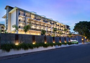 Un hôtel à plusieurs étages, doté de balcons, d’une façade bien éclairée et d’un parvis aménagé.