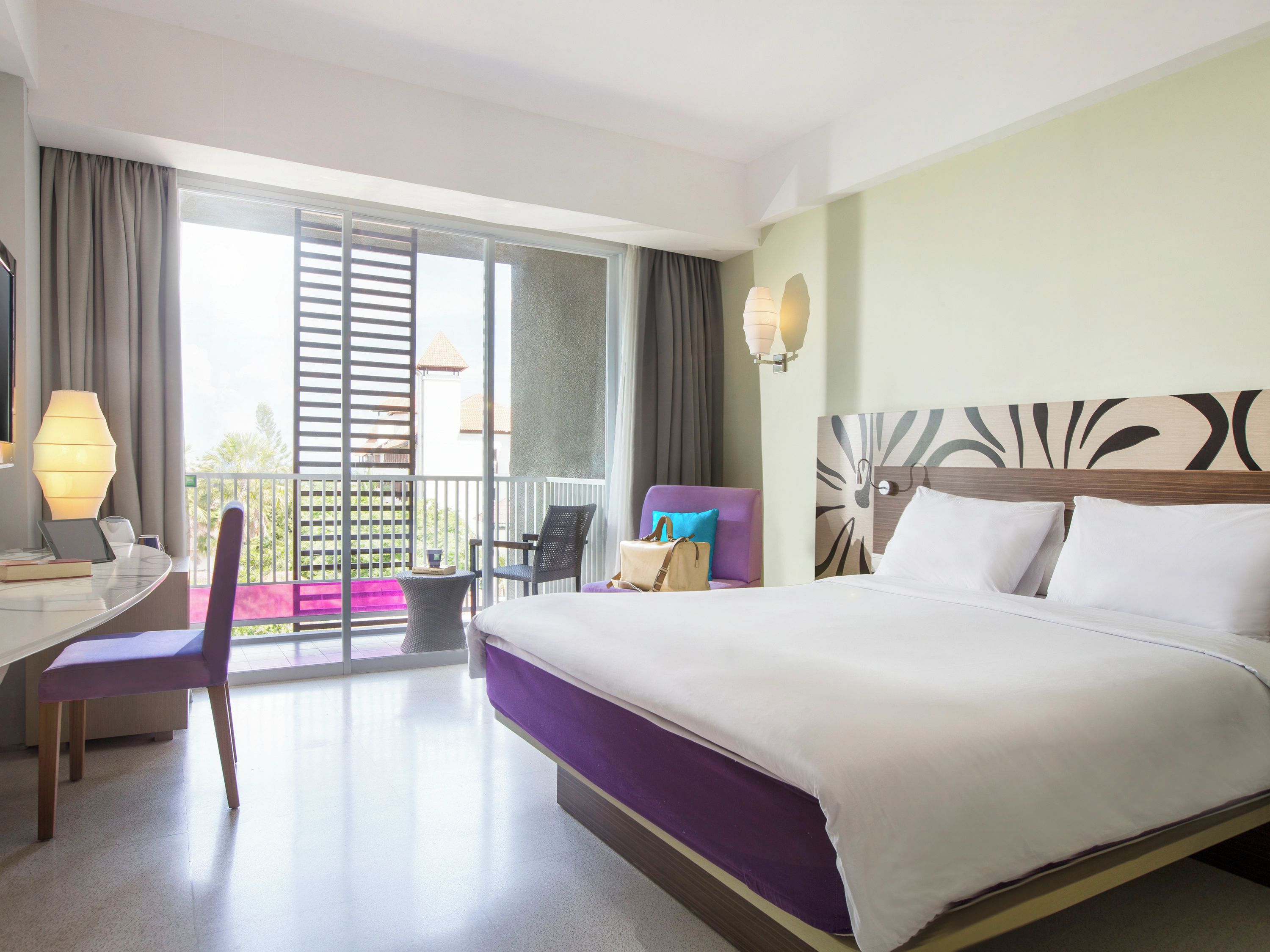 Quarto de hotel com cama, escrivaninha, cadeira, varanda e vista, além de parede com cabeceira decorativa.