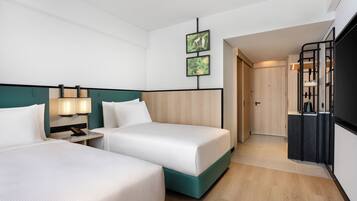 A hotel room with two beds, a desk with a lamp, a wardrobe, and a kitchenette.