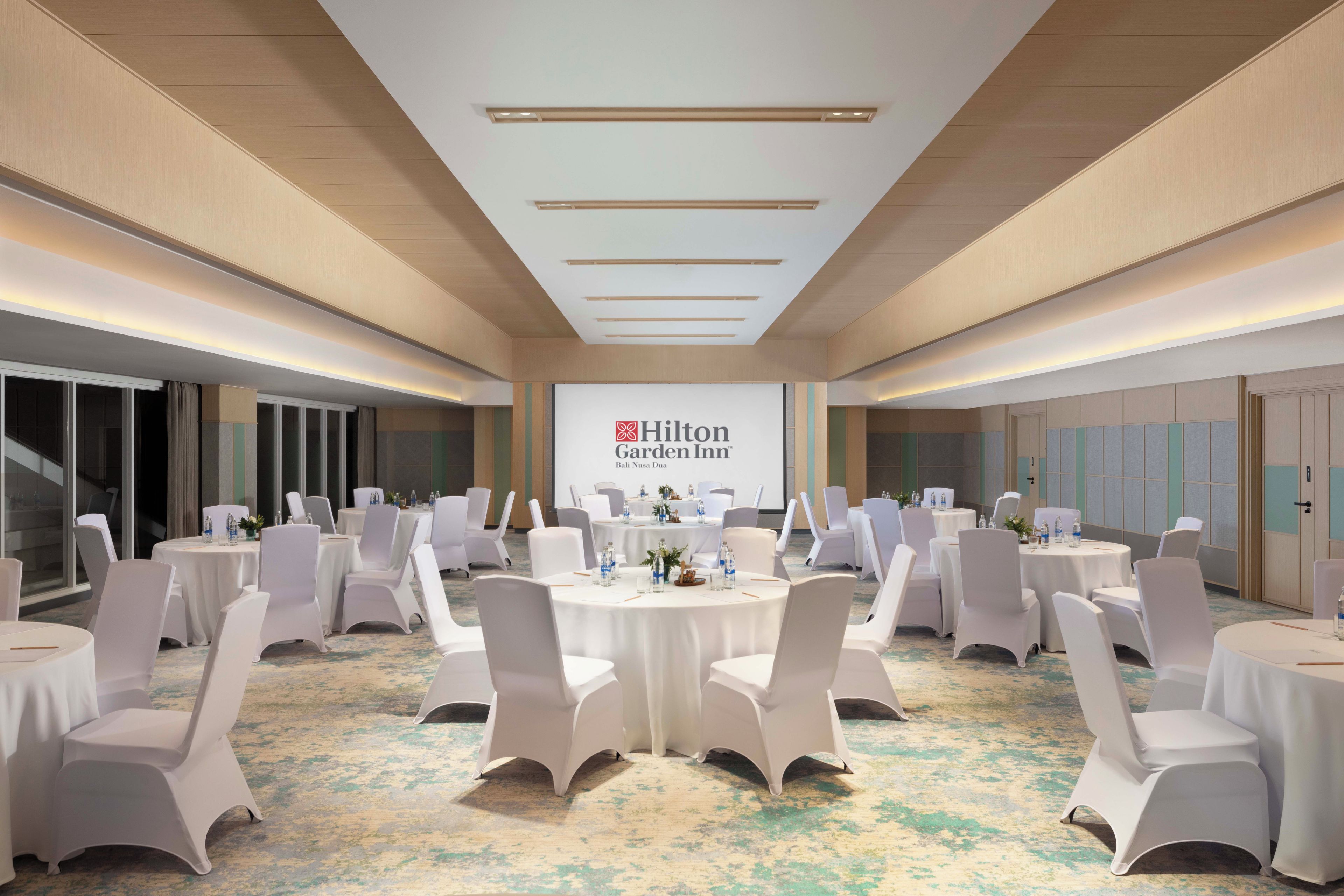 A conference room with round tables, white chairs, and a large screen displaying the Hilton Garden Inn logo.
