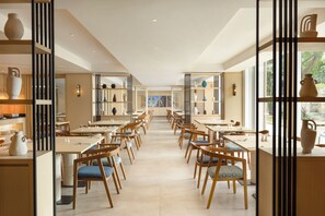 A spacious restaurant with multiple tables and chairs, wooden shelves with decorative items, and large windows allowing natural light. - Hilton Garden Inn Bali Nusa Dua (Nusa Dua)