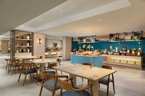 A modern dining area with wooden tables and chairs, a central buffet with various food items, and a blue accent wall. - Hilton Garden Inn Bali Nusa Dua (Nusa Dua)