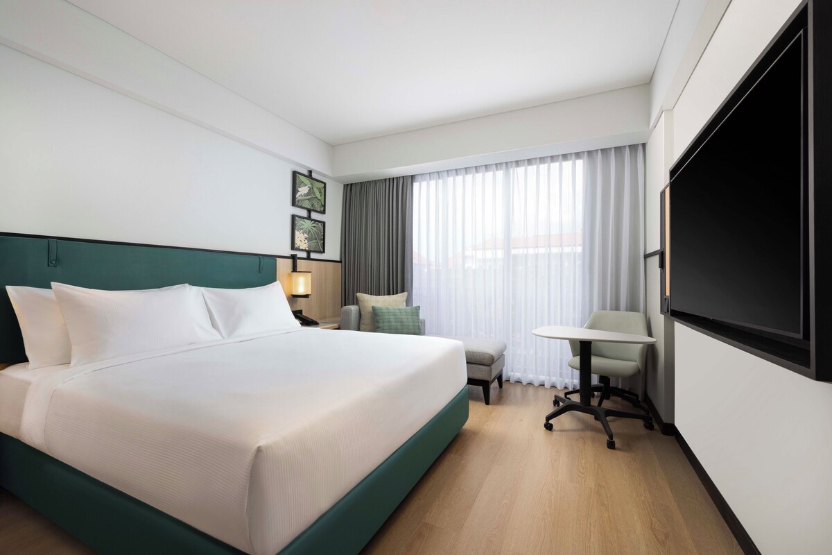 Ein Hotelzimmer mit einem großen Bett, einem Schreibtisch, einem Sessel, einem Fernseher und einem Fenster mit Vorhängen.