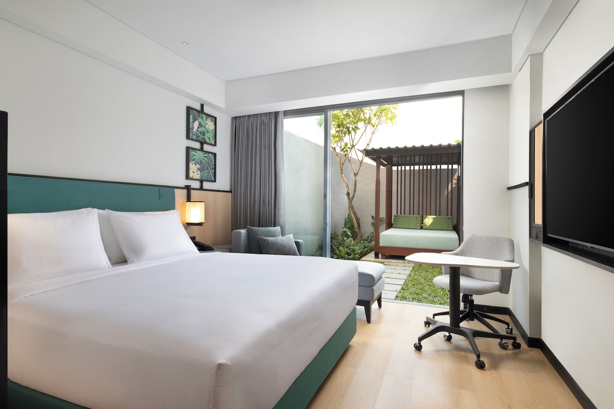 Ein modernes Hotelzimmer mit einem großen Bett, einem Schreibtisch, einem Sessel und einem kleinen Garten, der durch die Schiebetüren zu sehen ist.