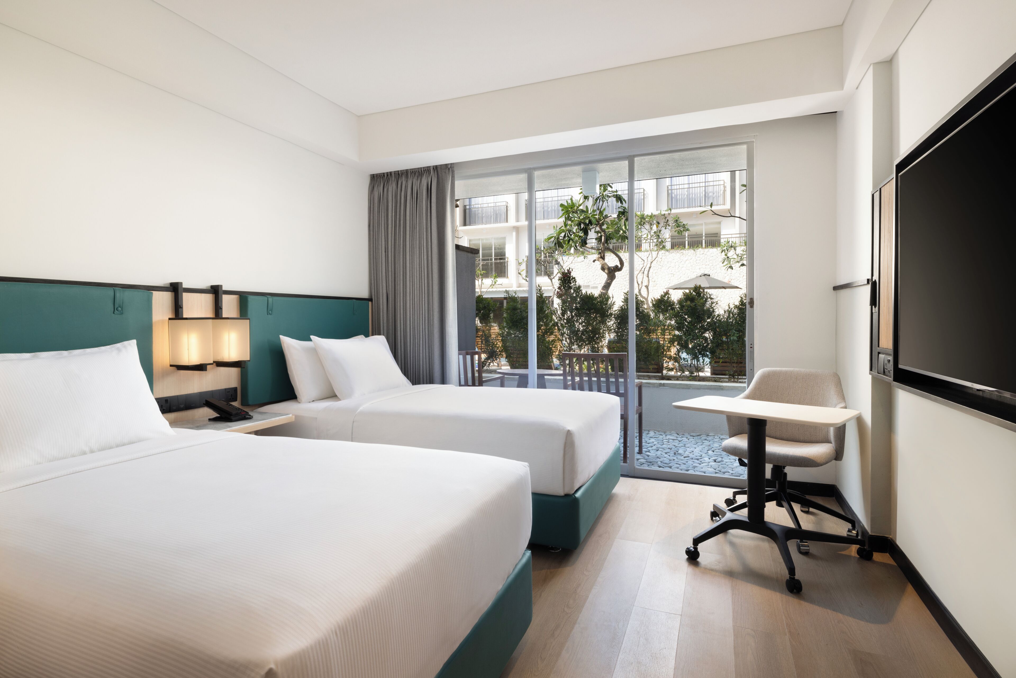 A hotel room with two beds, a desk, a chair, a TV, and a balcony with a view of the outside.