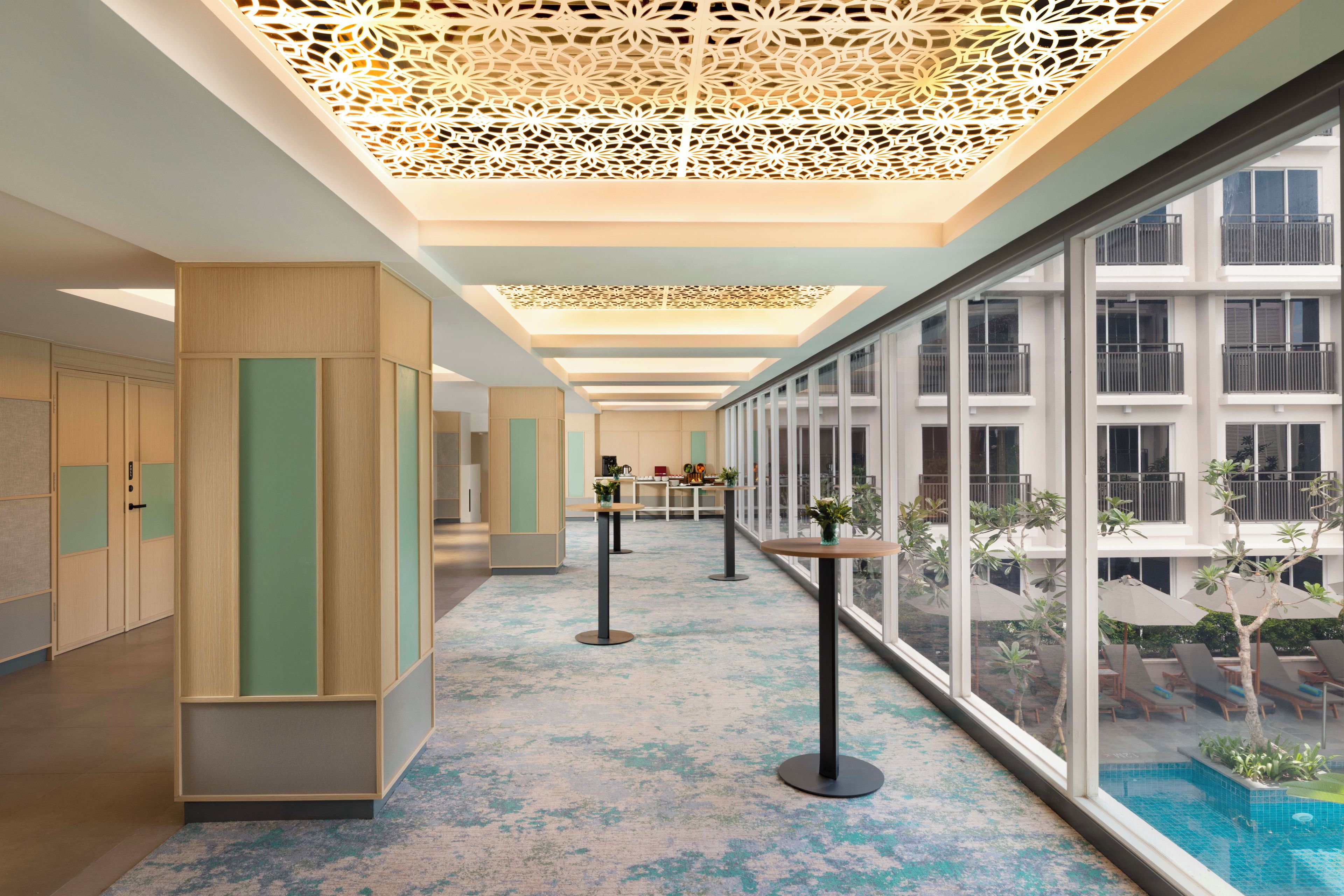 A modern hotel corridor with a pool view, wooden paneling, and a decorative ceiling.