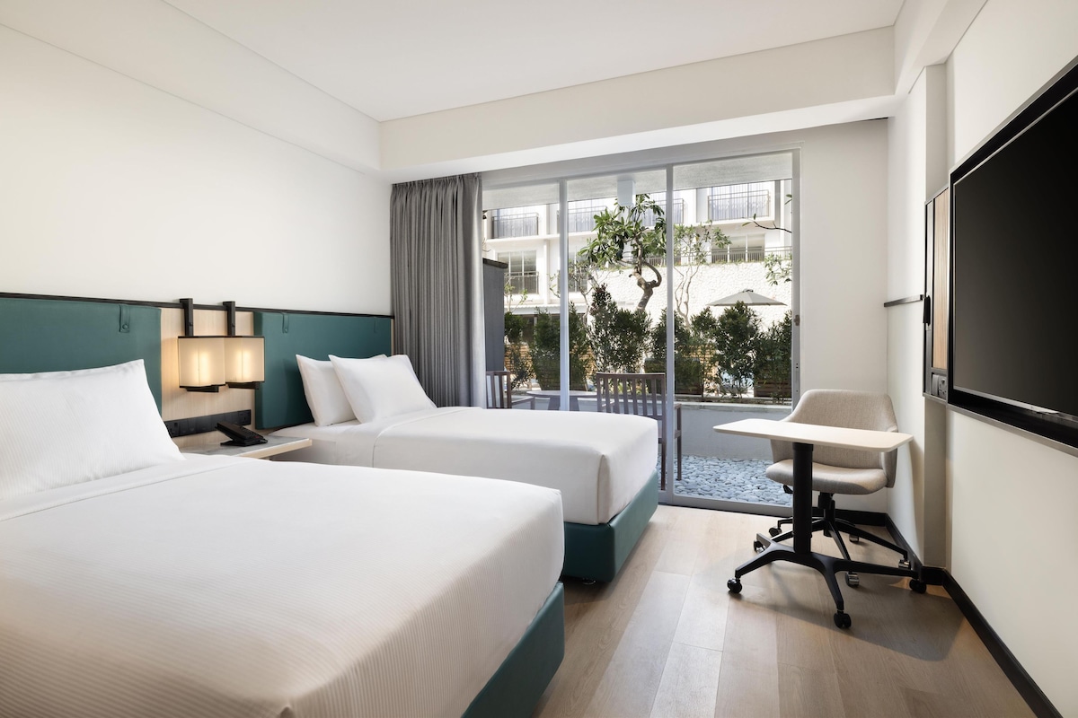 Ein Hotelzimmer mit zwei Betten, einem Schreibtisch, einem Stuhl, einem Fernseher und einem Balkon mit Blick.