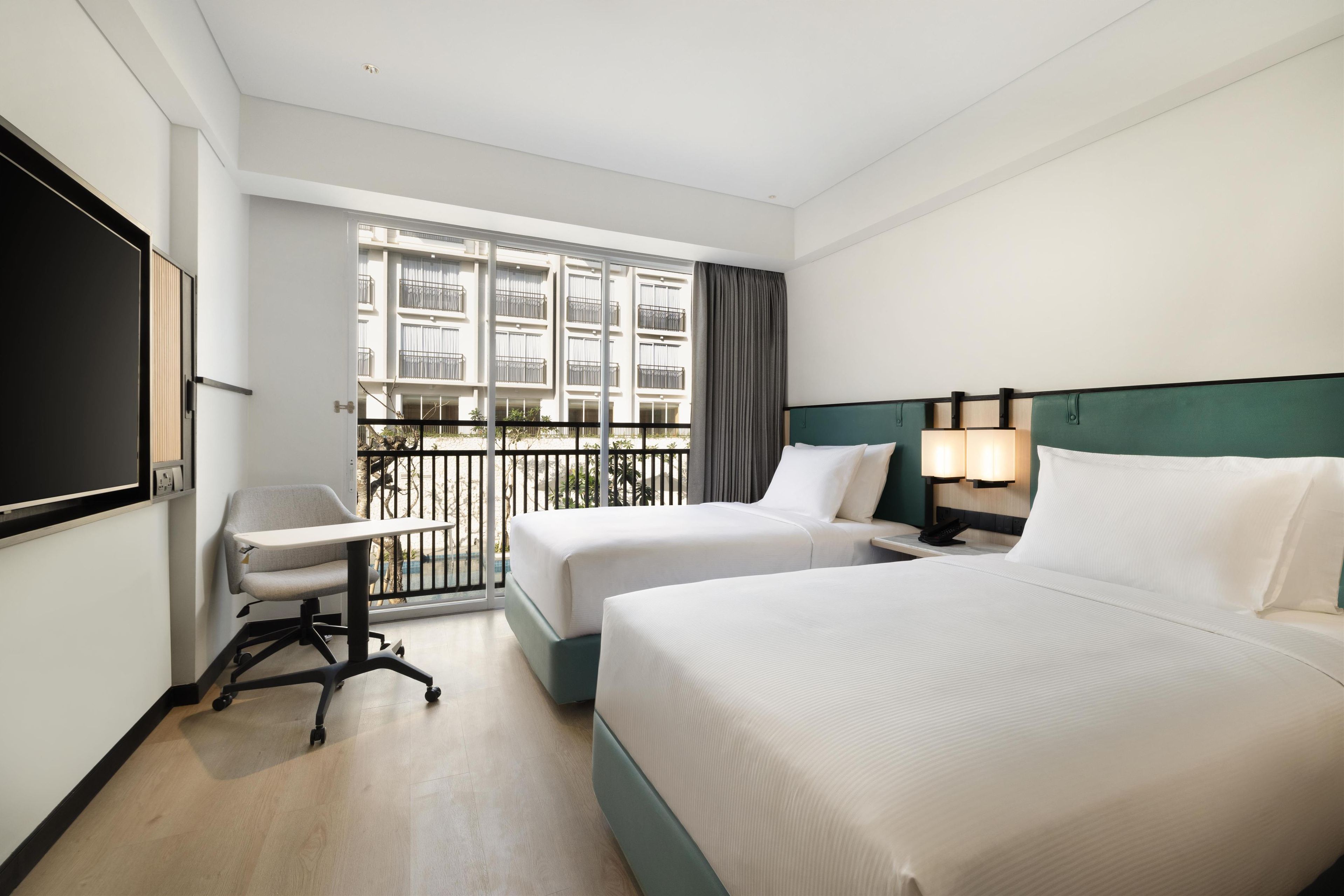 A hotel room with two beds, a desk, a chair, a TV, and a balcony with a view of other buildings.