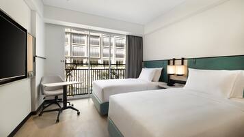 A hotel room with two beds, a desk, a chair, a TV, and a balcony with a view of other buildings.