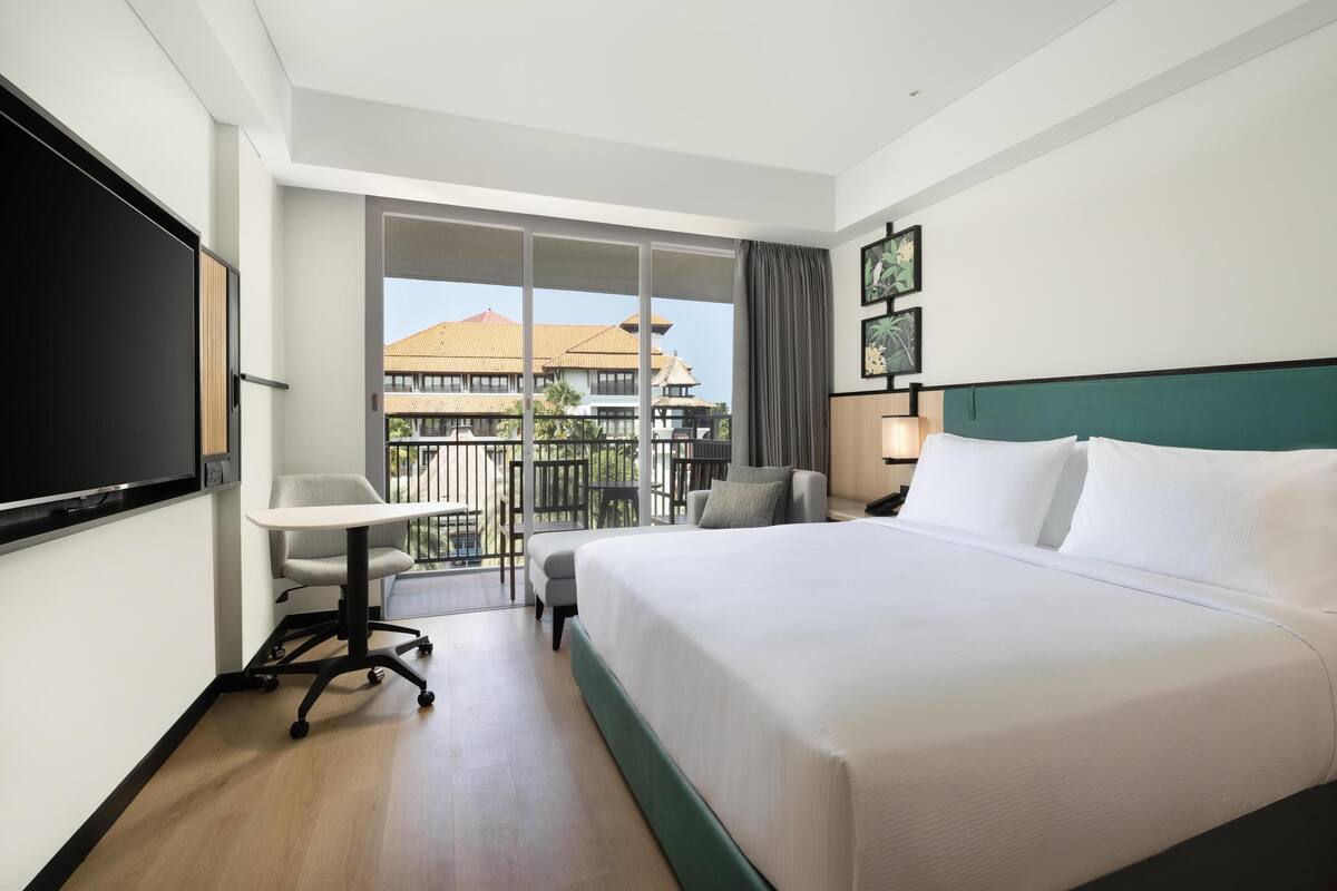 Ein Hotelzimmer mit Bett, Schreibtisch, Stuhl, Fernseher und einem Balkon mit Blick auf Gebäude.