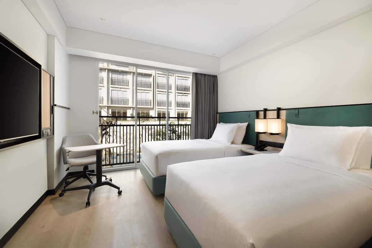 Ein Hotelzimmer mit zwei Betten, einem Schreibtisch, einem Sessel, einem Fernseher und einem Balkon mit Blick auf andere Gebäude.