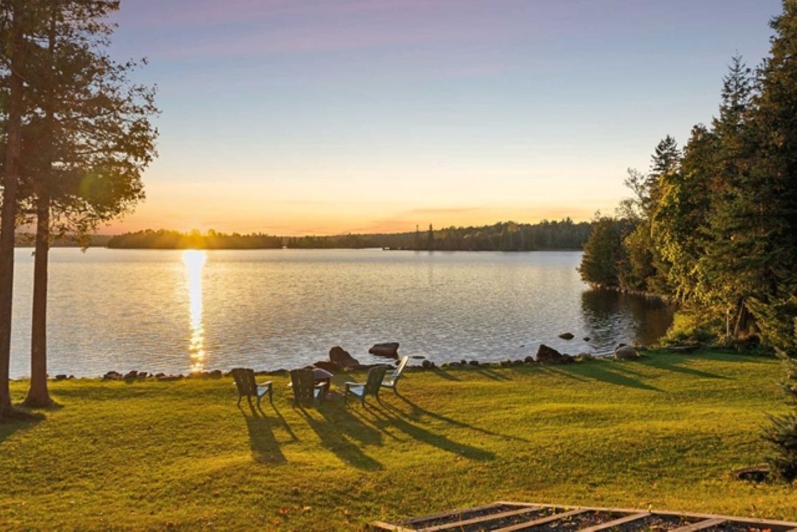 Vista sobre un lago con zona verde, una terraza de madera y una fila de sillas orientadas al agua.