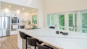 A modern kitchen with white cabinets, stainless steel appliances, a marble countertop, and large windows offering a view of greenery.
