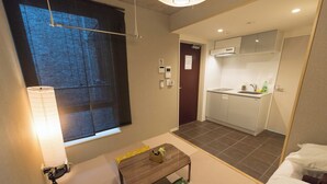 Une chambre d’hôtel compacte comprenant un coin cuisine, une petite table avec un coussin, une fenêtre avec un luminaire et une porte donnant accès à la salle de bain.