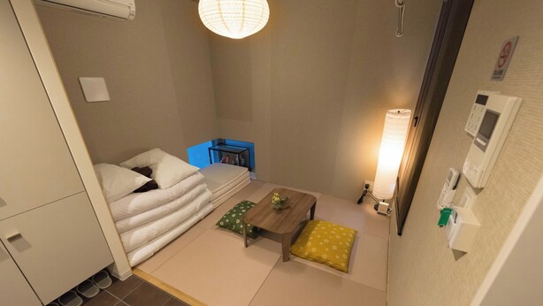 Une chambre d’hôtel de taille réduite, comprenant un lit, une petite table, une lampe et une poubelle.