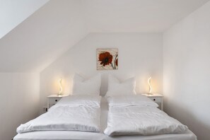 A bedroom with a sloped ceiling, two bedside tables with lamps, a bed with white bedding, and a painting on the wall.