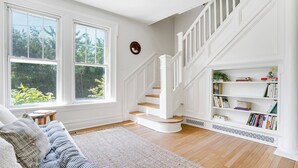 Amplia sala de estar con escalera, ventanas grandes, estantería y un rincón de descanso acogedor.