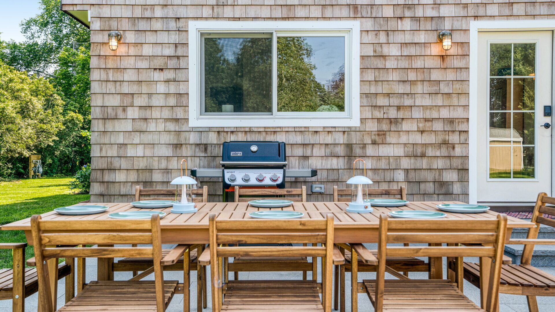 An outdoor dining area with a wooden table and chairs, a grill, and a window overlooking a garden.