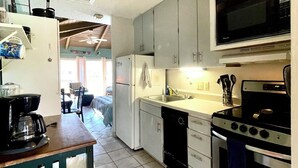 A compact kitchen with a refrigerator, microwave, stove, and sink. There is a coffee maker on the counter and a dishwasher. The kitchen has light-colored cabinets and a tiled floor. - Charming Studio condo in peaceful Kihei with AC, WiFi, pool (Kihei)