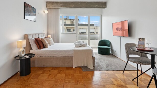 A modern hotel room with a bed, a desk, a chair, a TV, and a window with a city view. - GuestReady – A welcoming space to unwind (Porto)