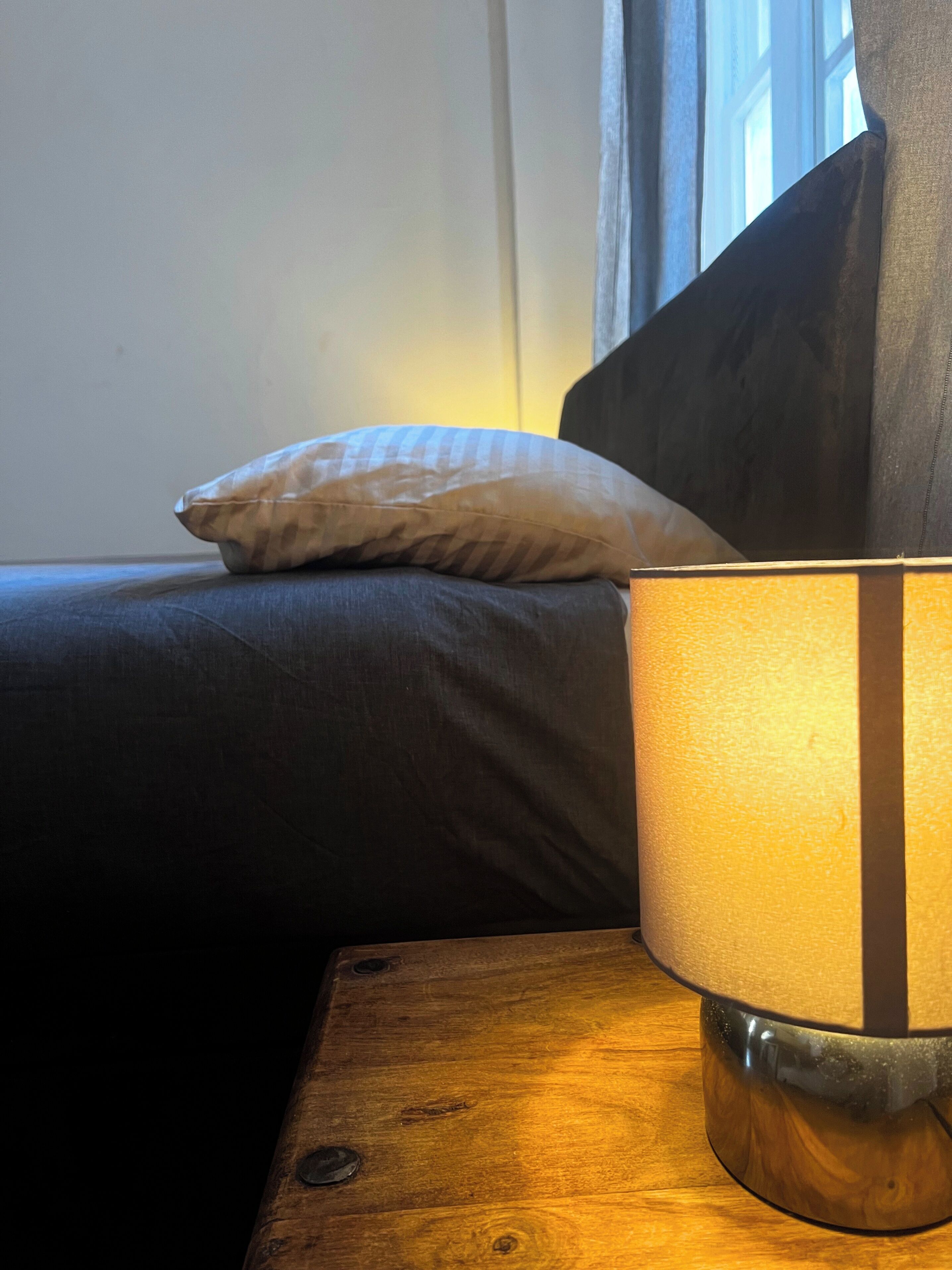 Una lámpara sobre una mesita de noche de madera al lado de una cama con un almohadón.
