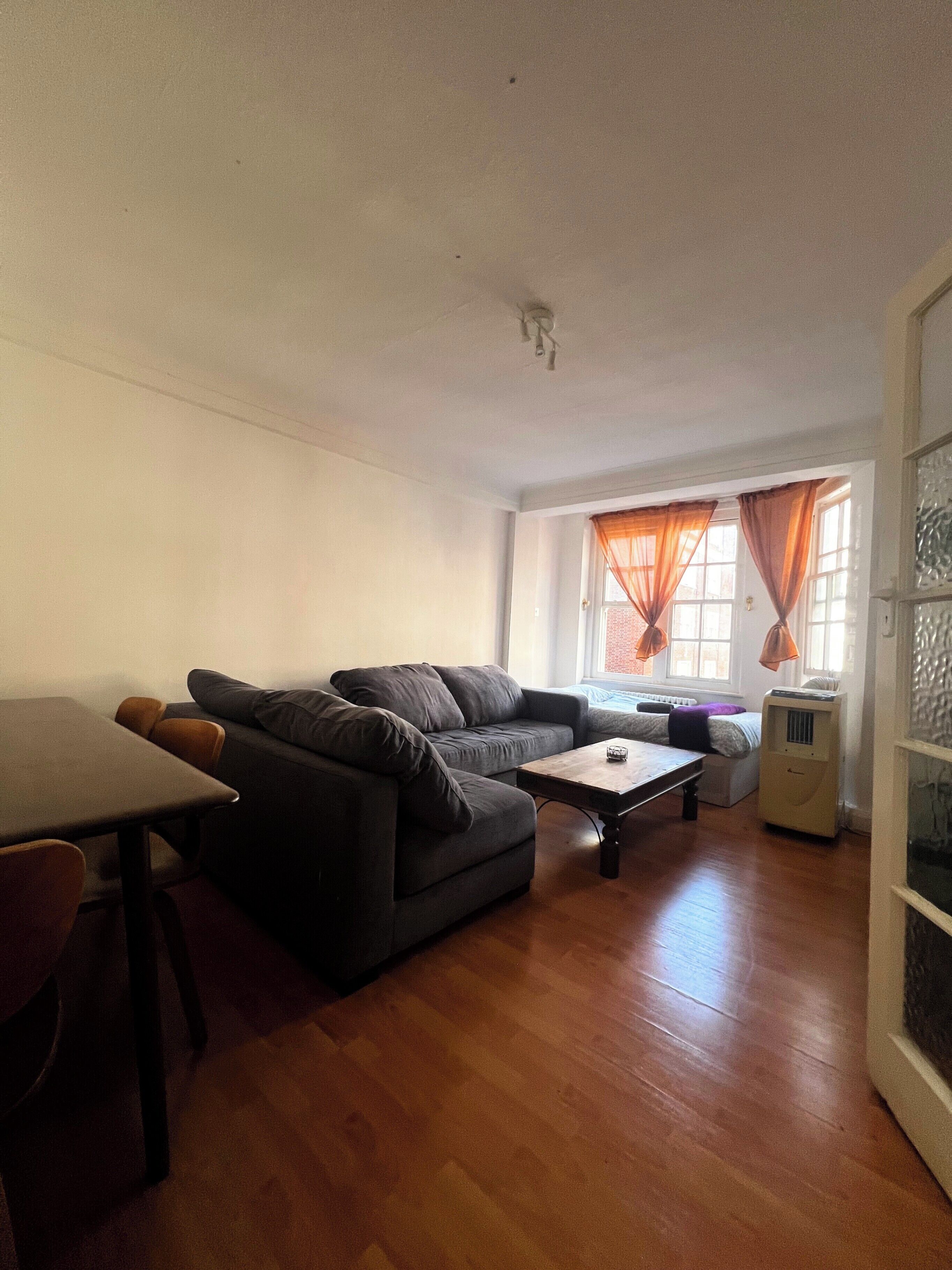 Een woonkamer met een bank, een salontafel en een eettafel. Er is een raam met gordijnen en een radiator.