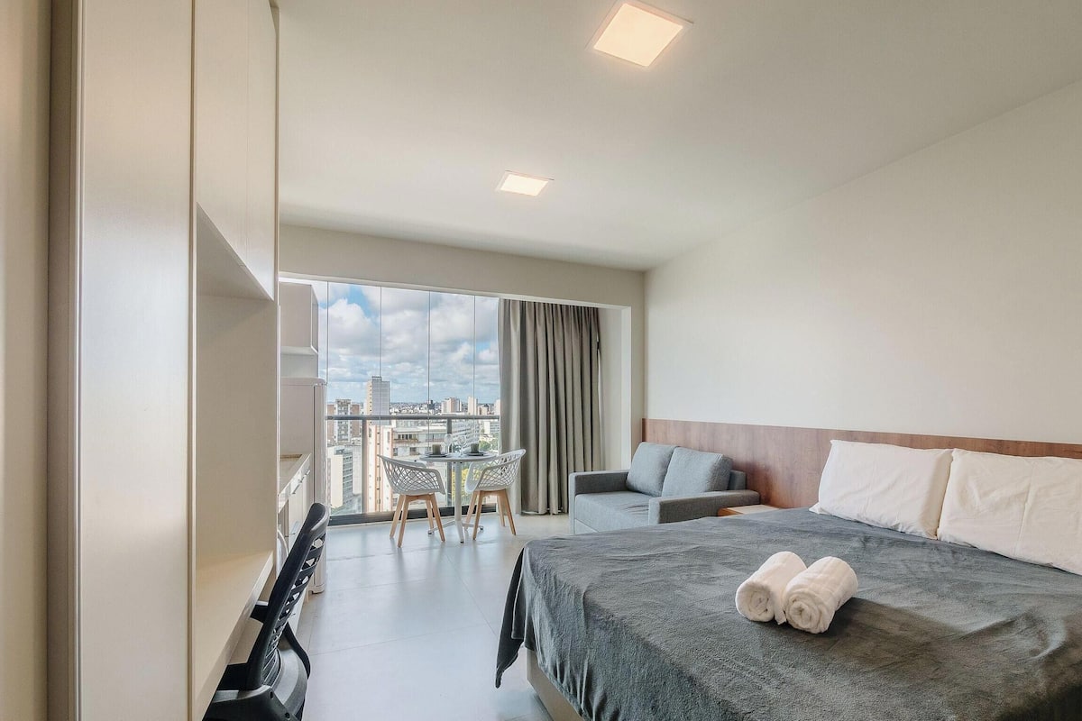 A hotel room with a bed, a chair, a table, and a balcony with a city view.